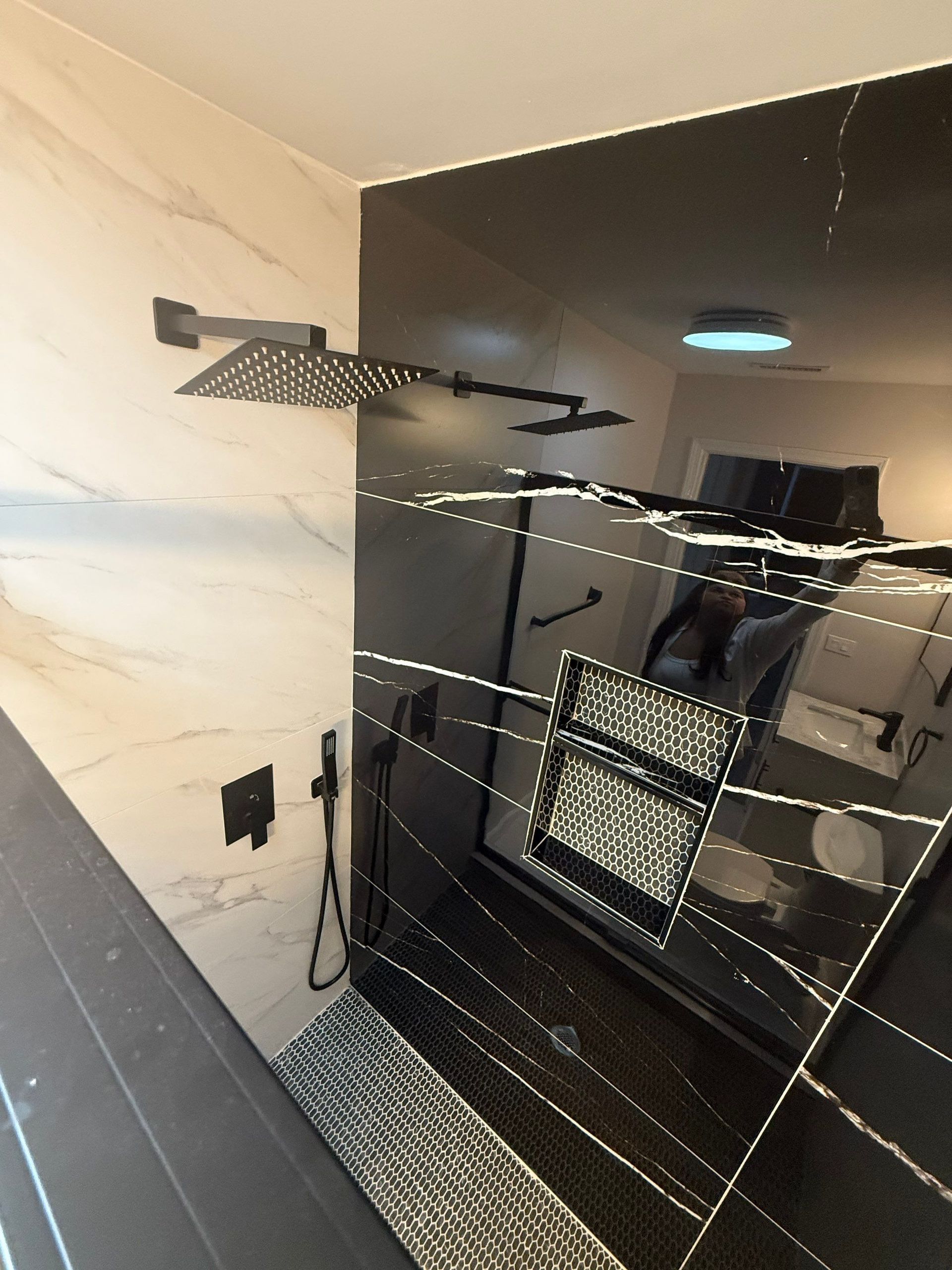 Shower with black and white tile walls, a rain shower head, and a mirrored wall reflecting a room.