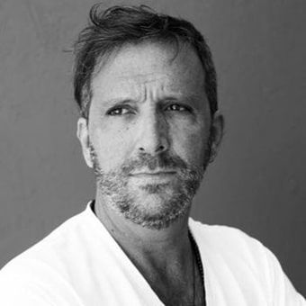 A black and white photo of a man with a beard wearing a white scrub top.