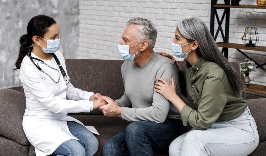 A doctor is sitting on a couch with a man and woman wearing face masks.