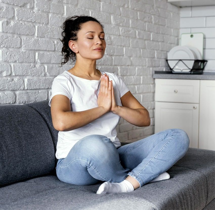 A woman is sitting on a couch with her eyes closed and her hands folded in prayer.