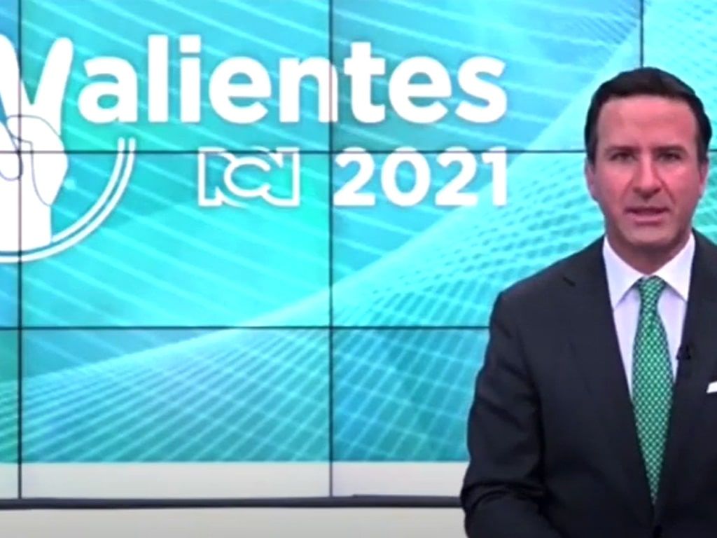 A man in a suit and tie stands in front of a wall that says valientes 2021