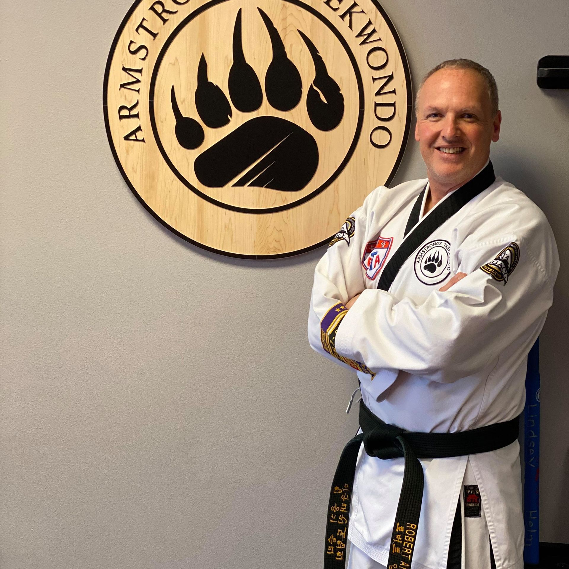 Man in black belt and TaeKwonDo uniform smiles, standing near a wooden logo with a bear paw.