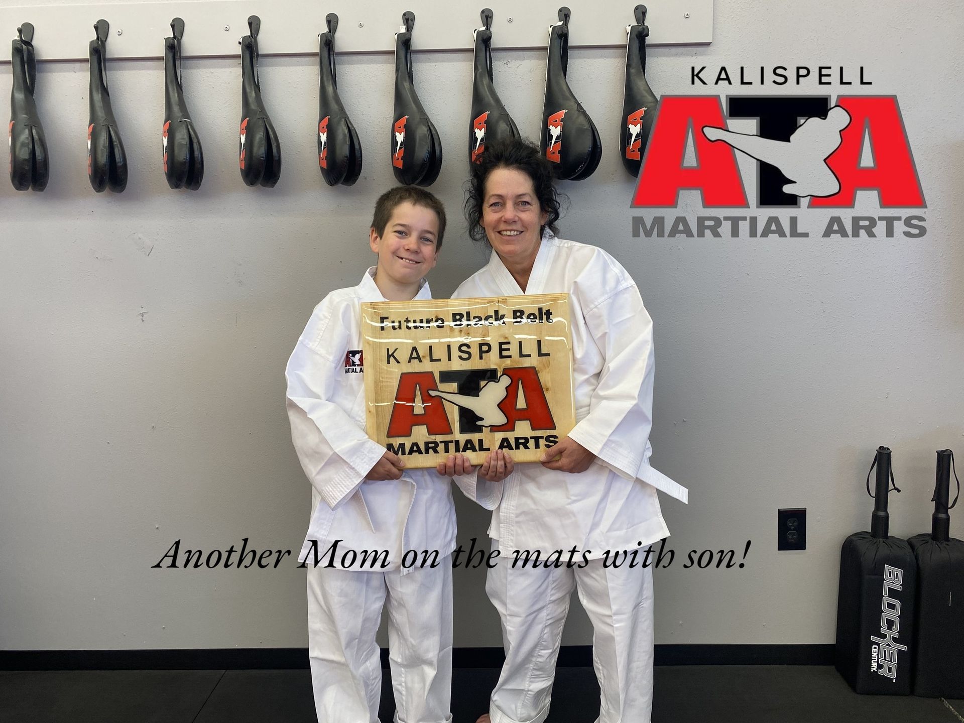 Mother and son in white martial arts uniforms hold a wooden sign with