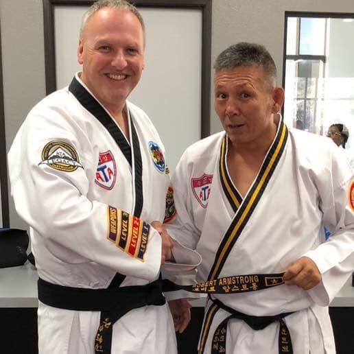Two men in martial arts uniforms, one shaking hands and holding a black belt with name on it.
