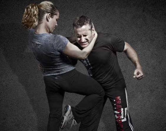 Woman in gray shirt and black pants kicking a man in a self-defense setting. Both display aggressive expressions.