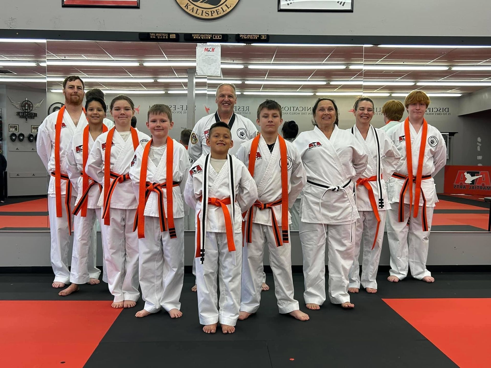 Group of people in white karate uniforms, orange belts, smiling. Studio setting with mirrors.