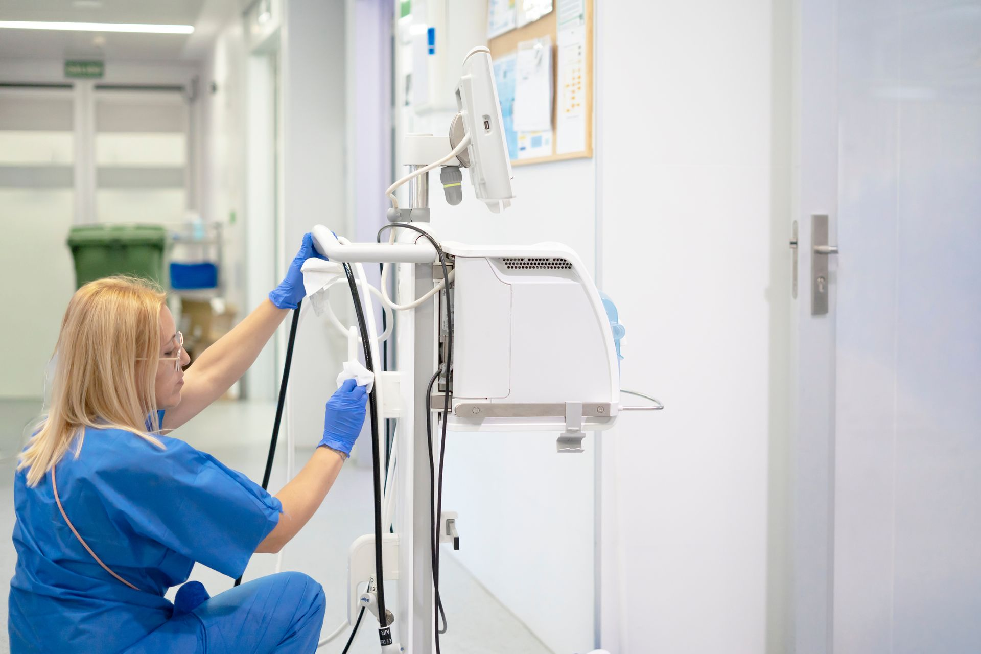 Medical worker in blue scrubs sanitizing a ventilator in a hospital hallway.