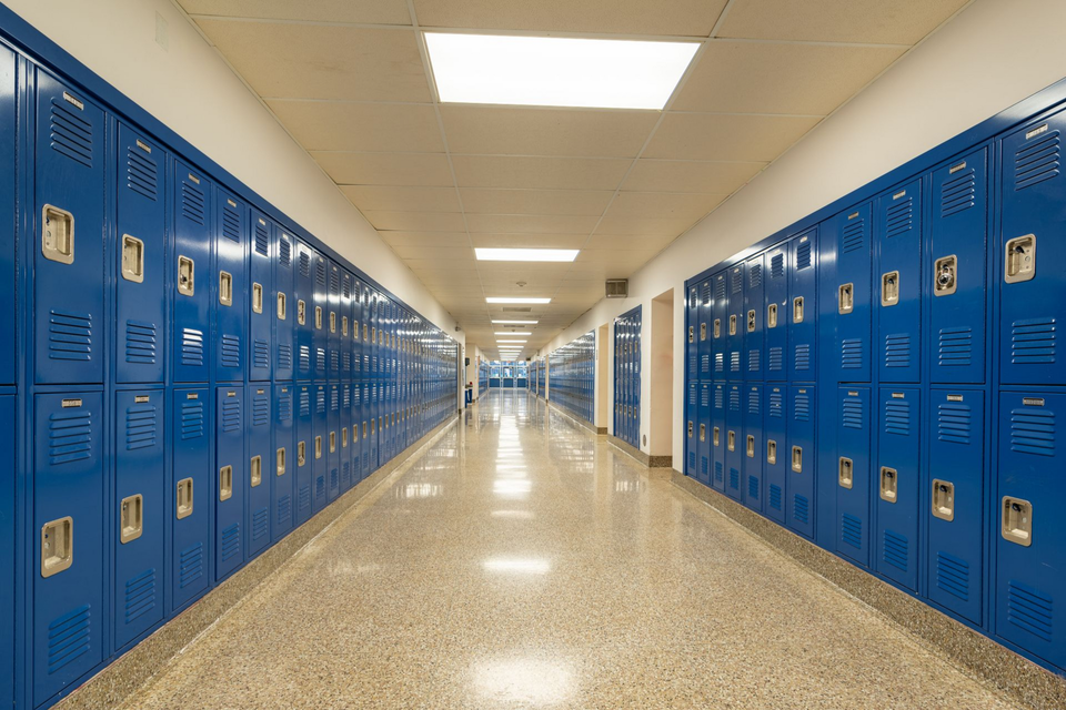 Blue school lockers line a long hallway with a speckled floor and bright overhead lights.