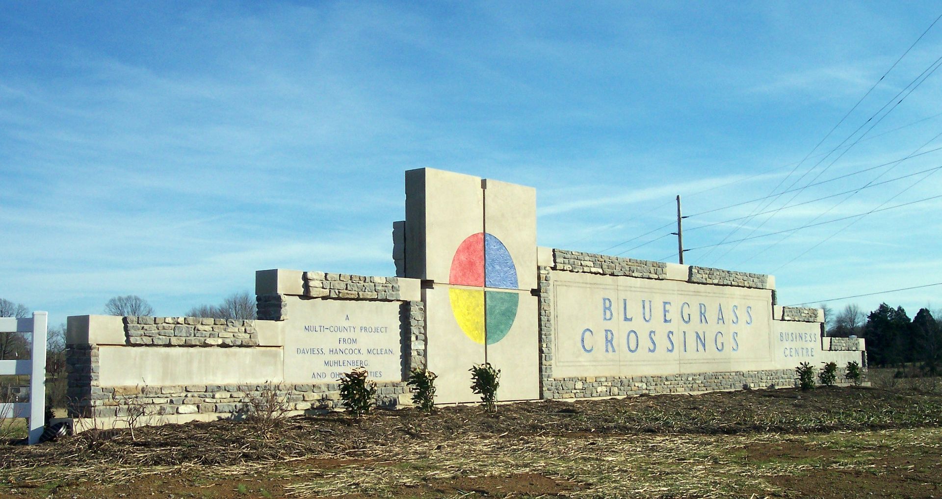 Stone roadside monument reading “BLEIBERG CROSSING” with a multicolored cross emblem under a blue sky