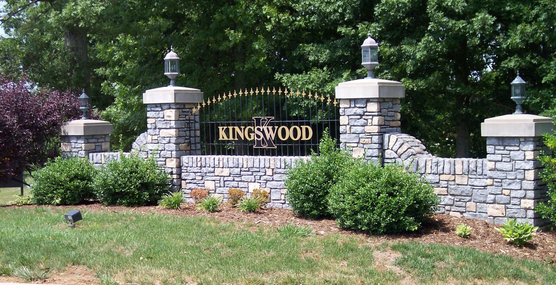 Stone entrance sign reading “Kingswood” amid landscaped shrubs and brick pillars at a residential community entrance