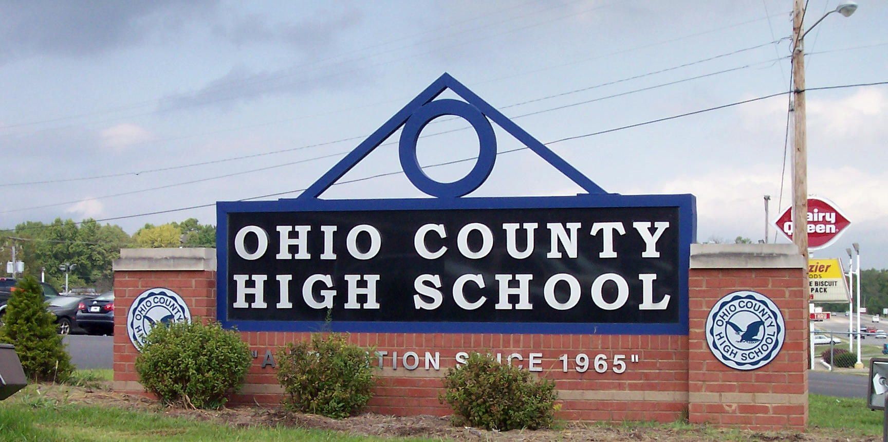 Ohio County High School sign with blue and white lettering on a brick base by a roadside.