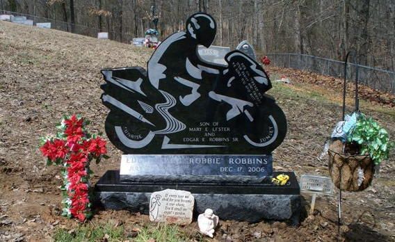 Motorcycle-shaped gravestone with flowers and a wreath in a wooded cemetery