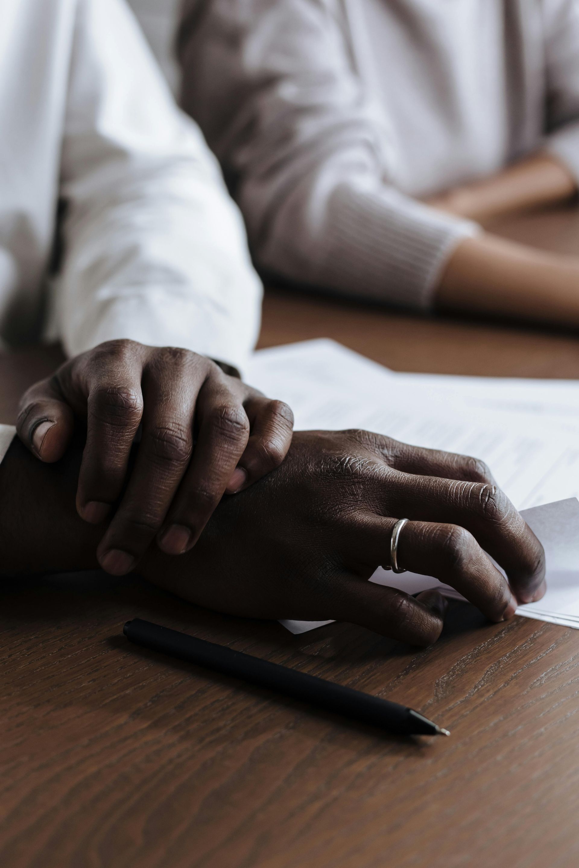 Two hands resting on papers at a wooden table with a pen nearby