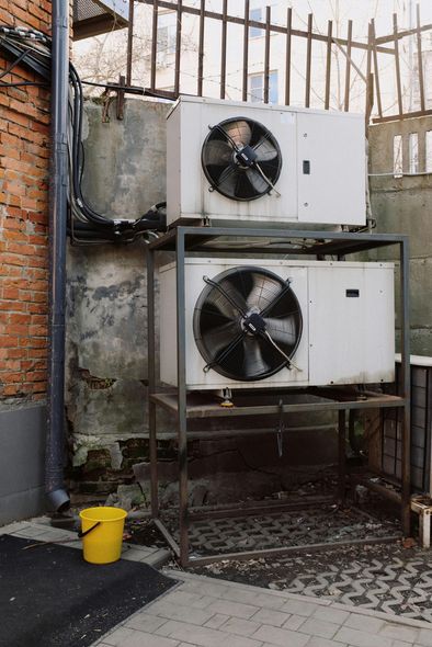 Two white air conditioning units stacked on a metal rack next to a brick wall. A yellow bucket sits below.