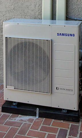 Samsung air conditioning unit, light-colored, against a wall on a brick patio.