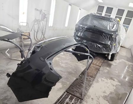 Car parts in a paint booth, ready for paint. Black fender in the foreground, car in background covered in plastic.