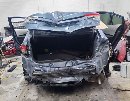 Heavily damaged blue car rear, trunk open, in a repair shop.