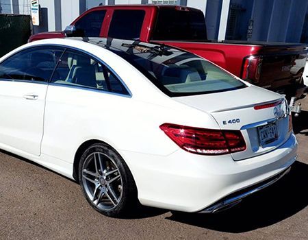 White Mercedes-Benz E 400 coupe parked, with a black roof, and red pickup truck behind.