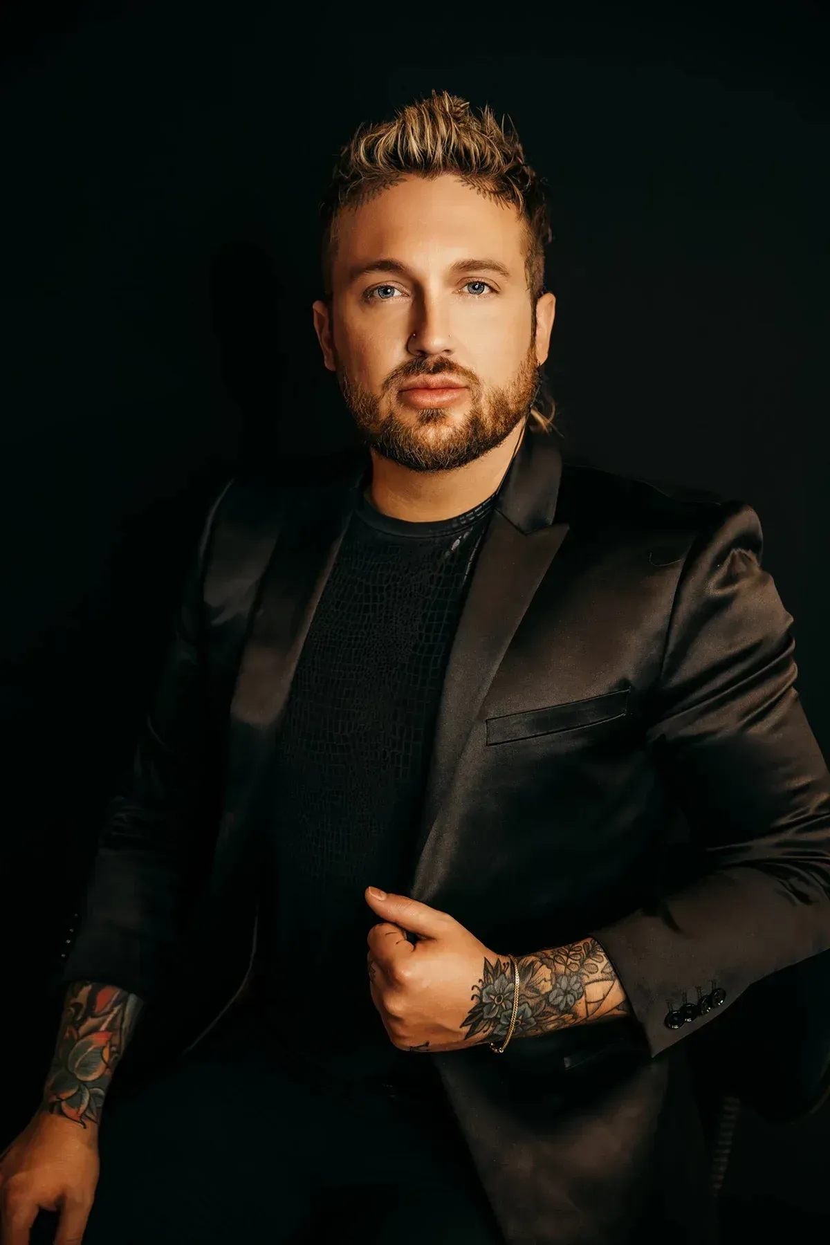 A man with blonde hair and tattoos poses in a black blazer. He is looking at the camera against a dark background.