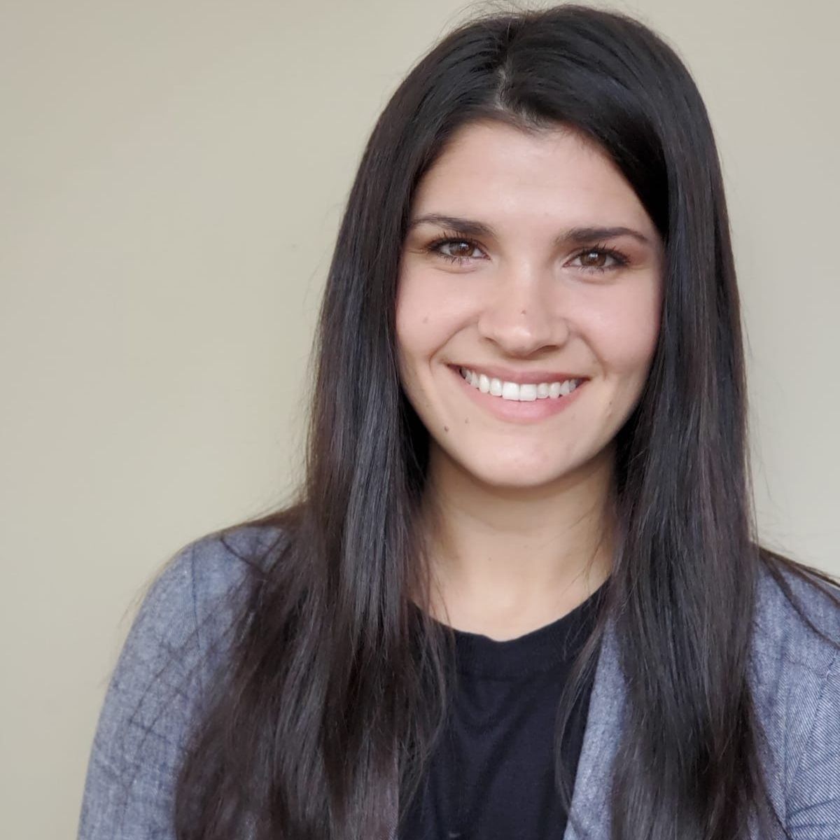 Woman with long dark hair smiles at the camera, wearing a grey blazer over a black shirt, against a neutral background.