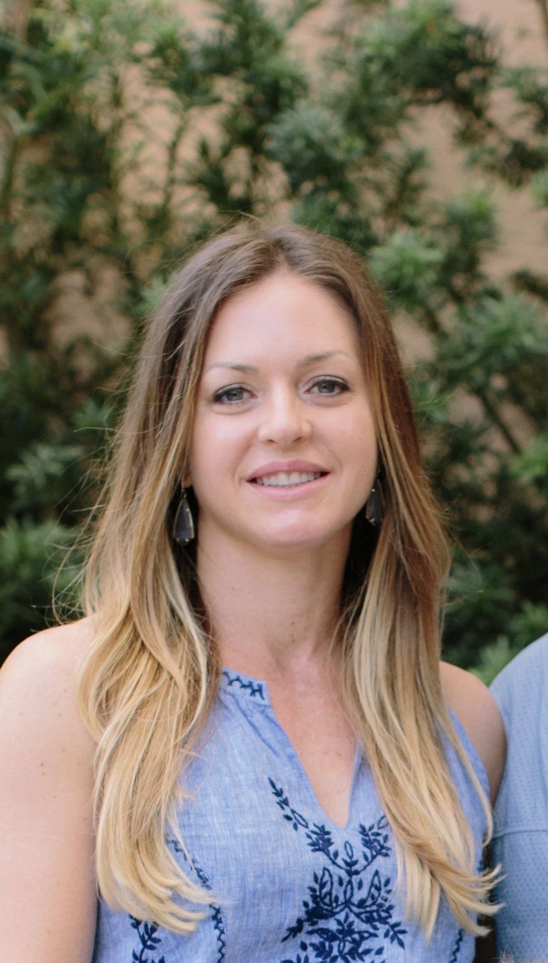 Woman with long, ombre blonde hair smiles in a light blue patterned top against a green leafy background.
