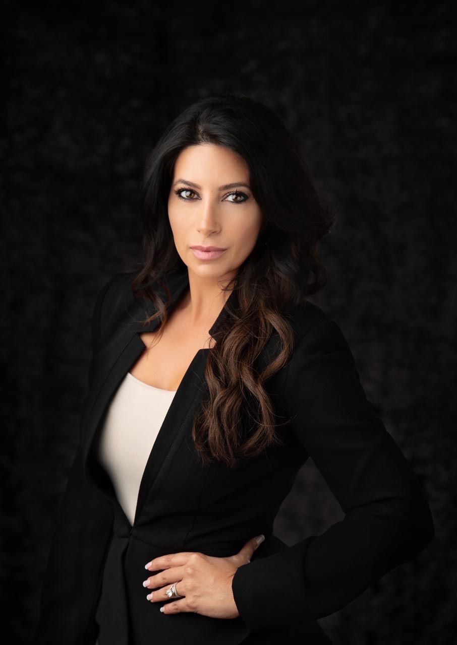 Woman in black blazer and white top poses confidently against a dark backdrop. She has long, dark hair and a serious expression.