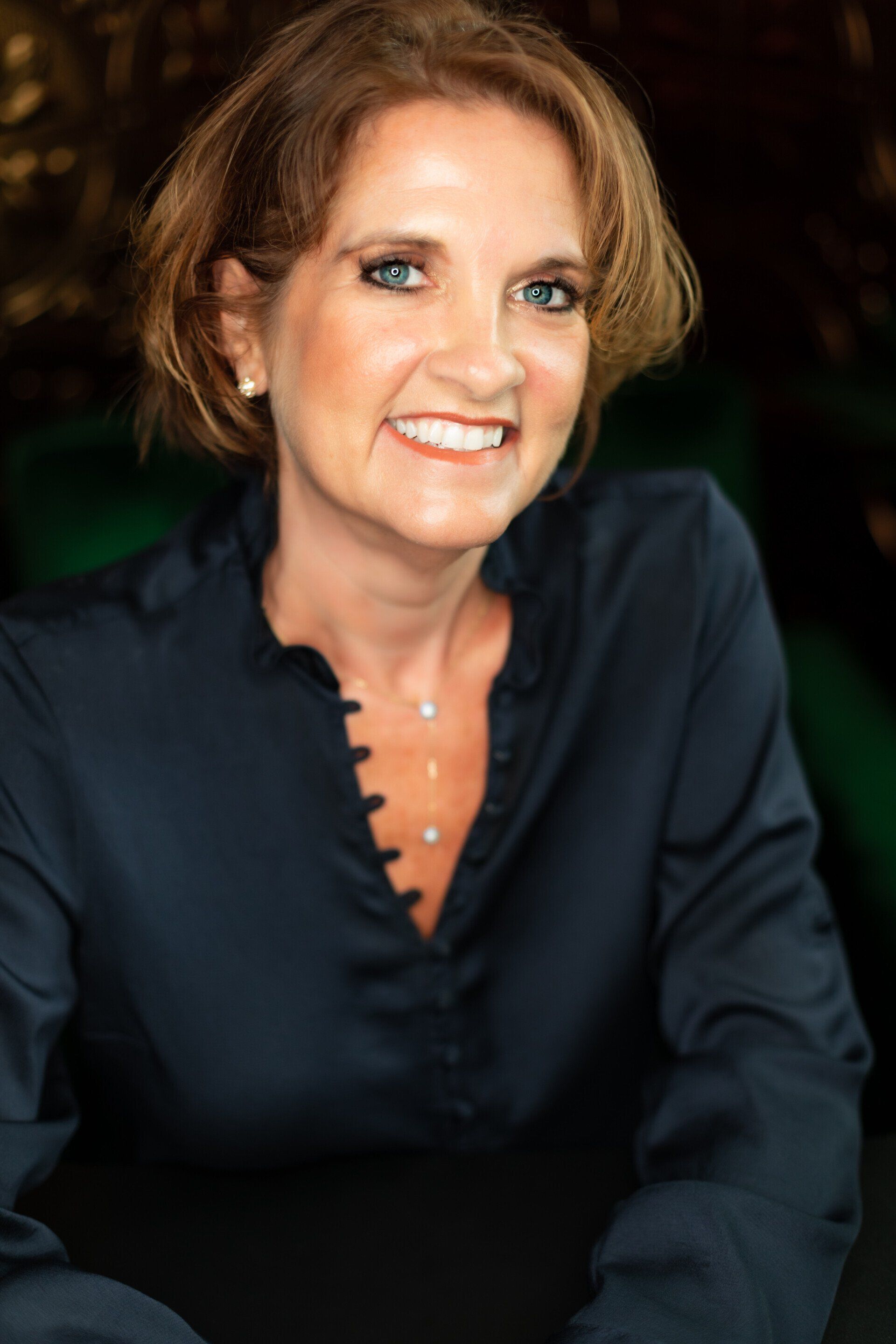 Woman with short brown hair, smiling, wearing a dark blue silk blouse, and a small necklace; dark background.