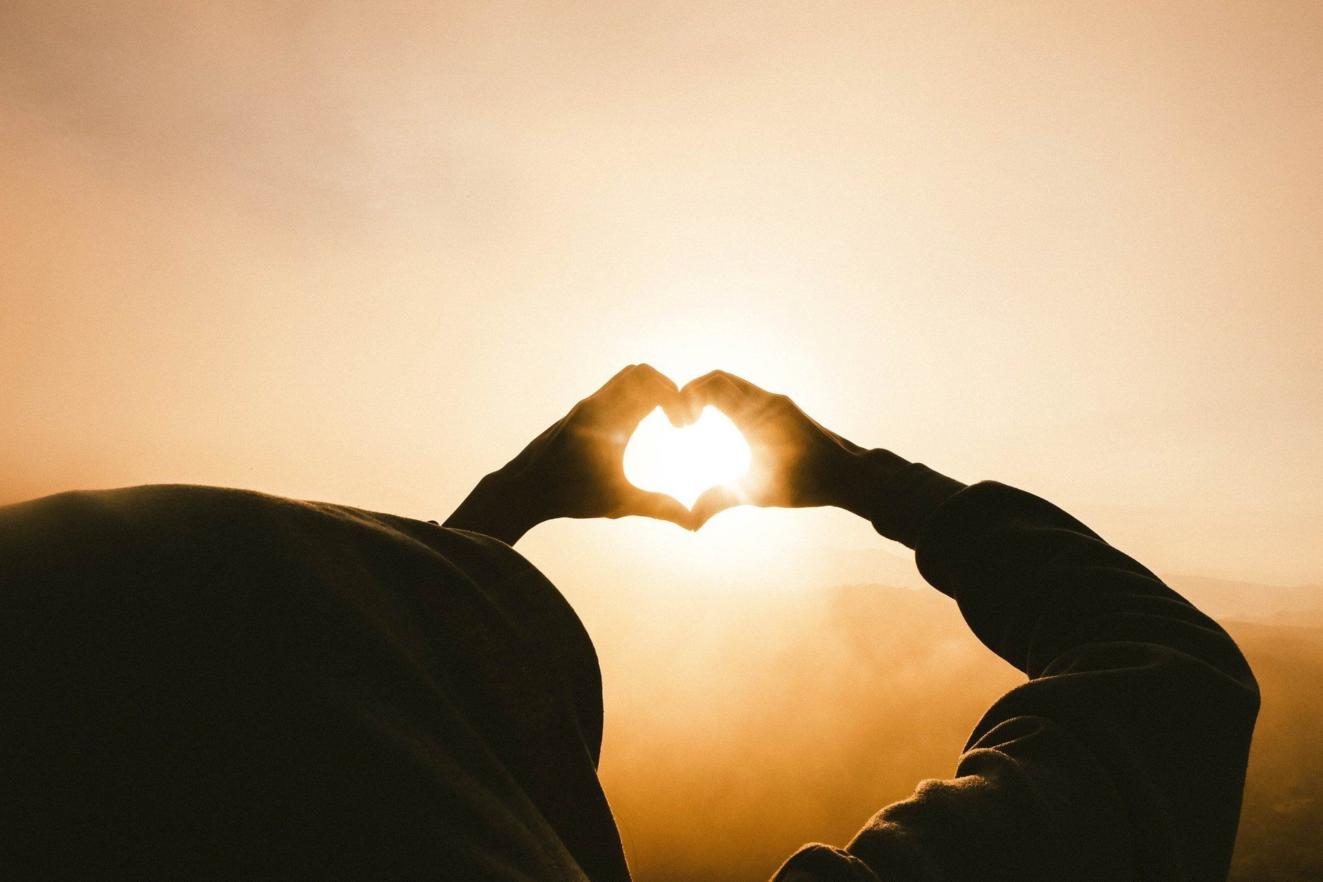 Silhouette of hands forming a heart around the setting sun, creating a warm glow in an open sky.