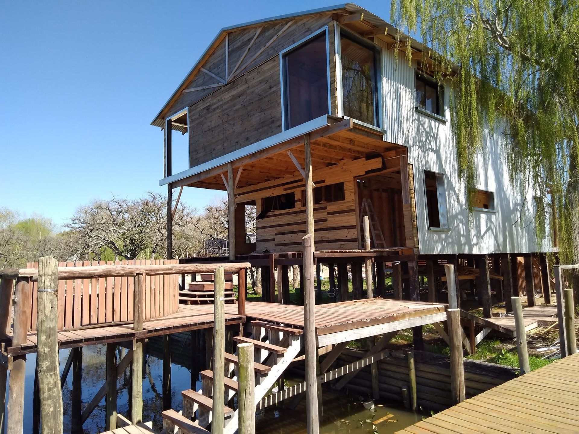 Casa de madera desgastada sobre pilotes junto al agua con terraza y rampa bajo un cielo azul claro.
