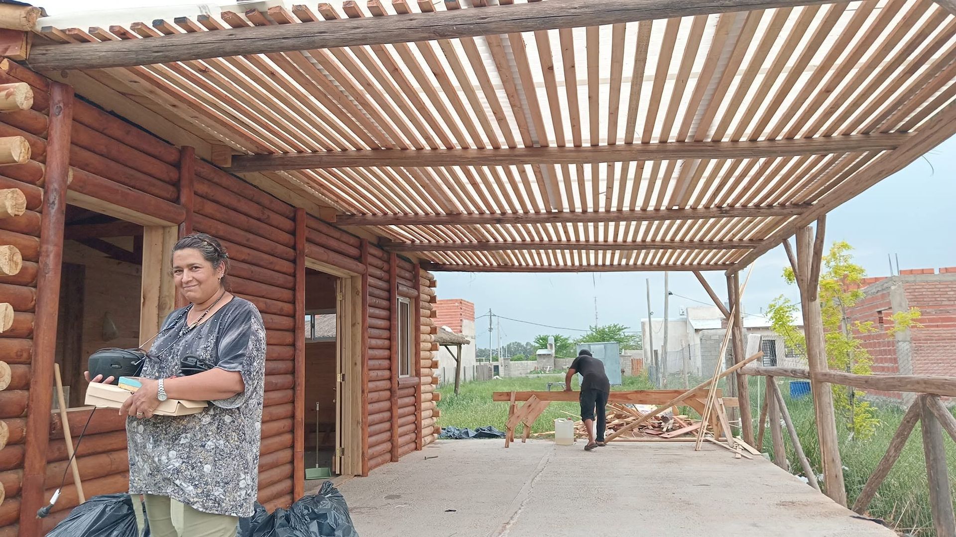 Patio exterior cubierto con techo de madera, un hombre de pie a la izquierda y otra persona al fondo cerca de una valla.