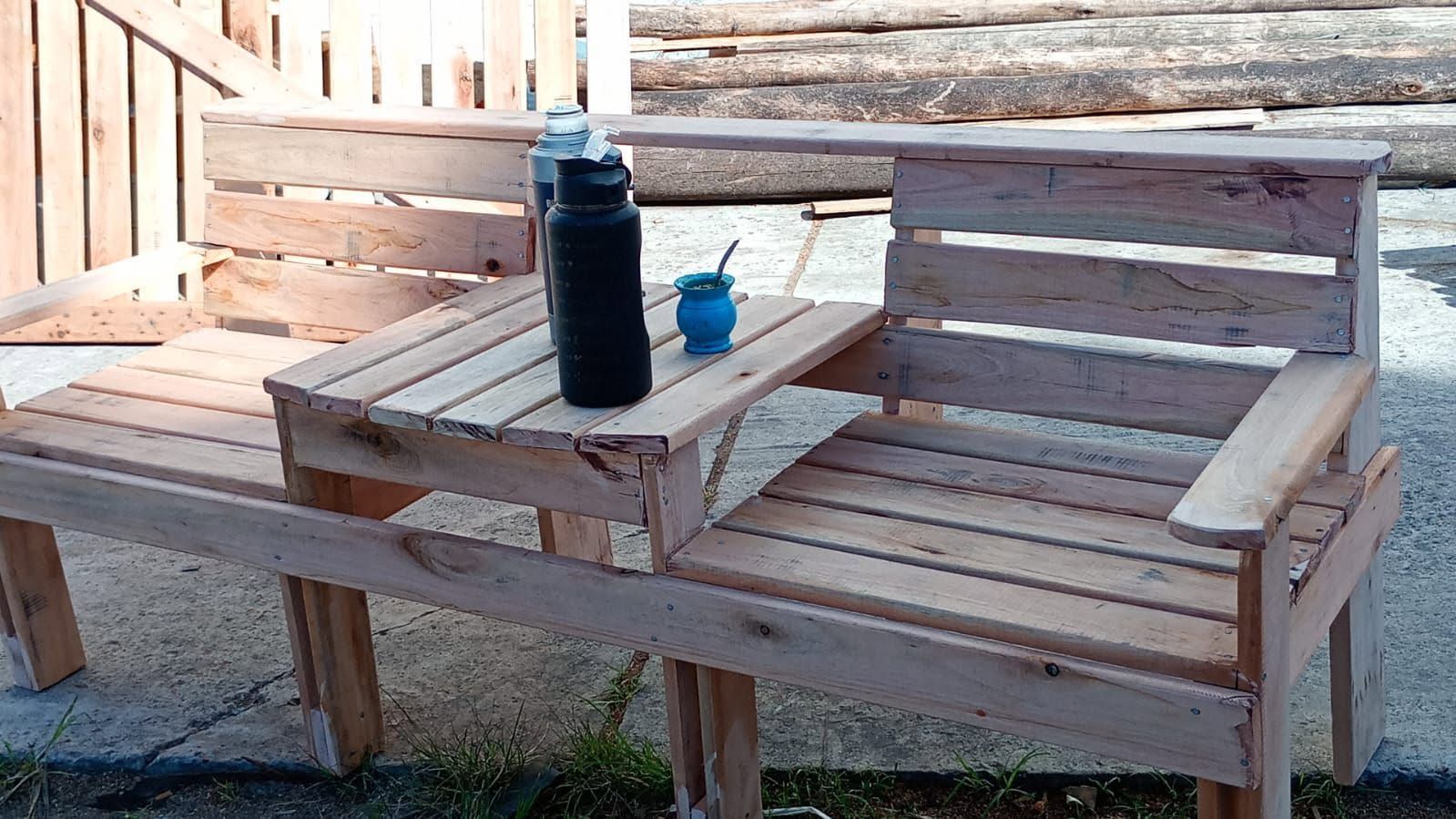Banco de madera para exteriores con mesa incorporada, que sostiene una botella negra y una taza azul
