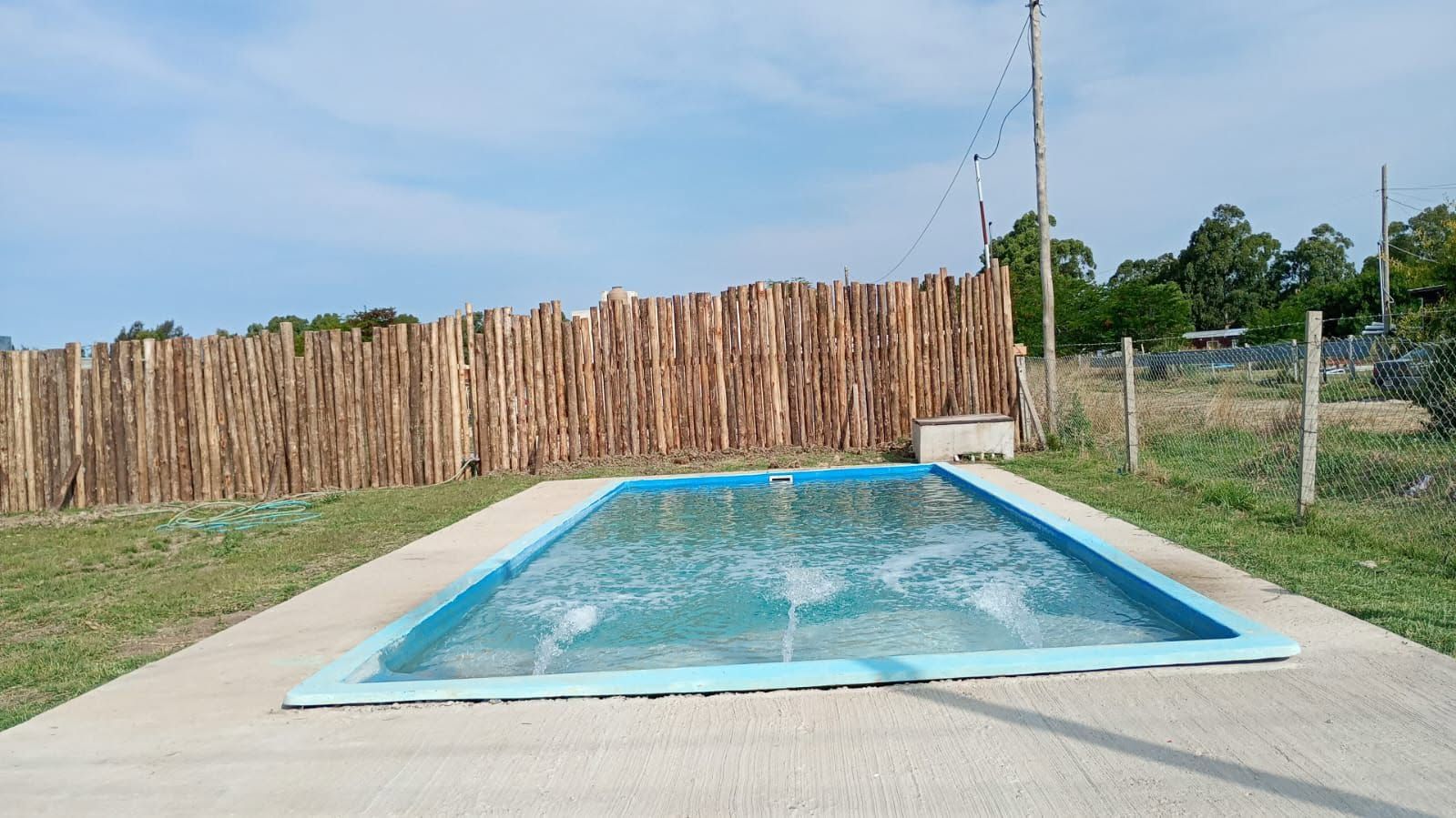 Piscina exterior vacía con agua azul, rodeada por una terraza de hormigón y una valla de madera bajo un cielo despejado.