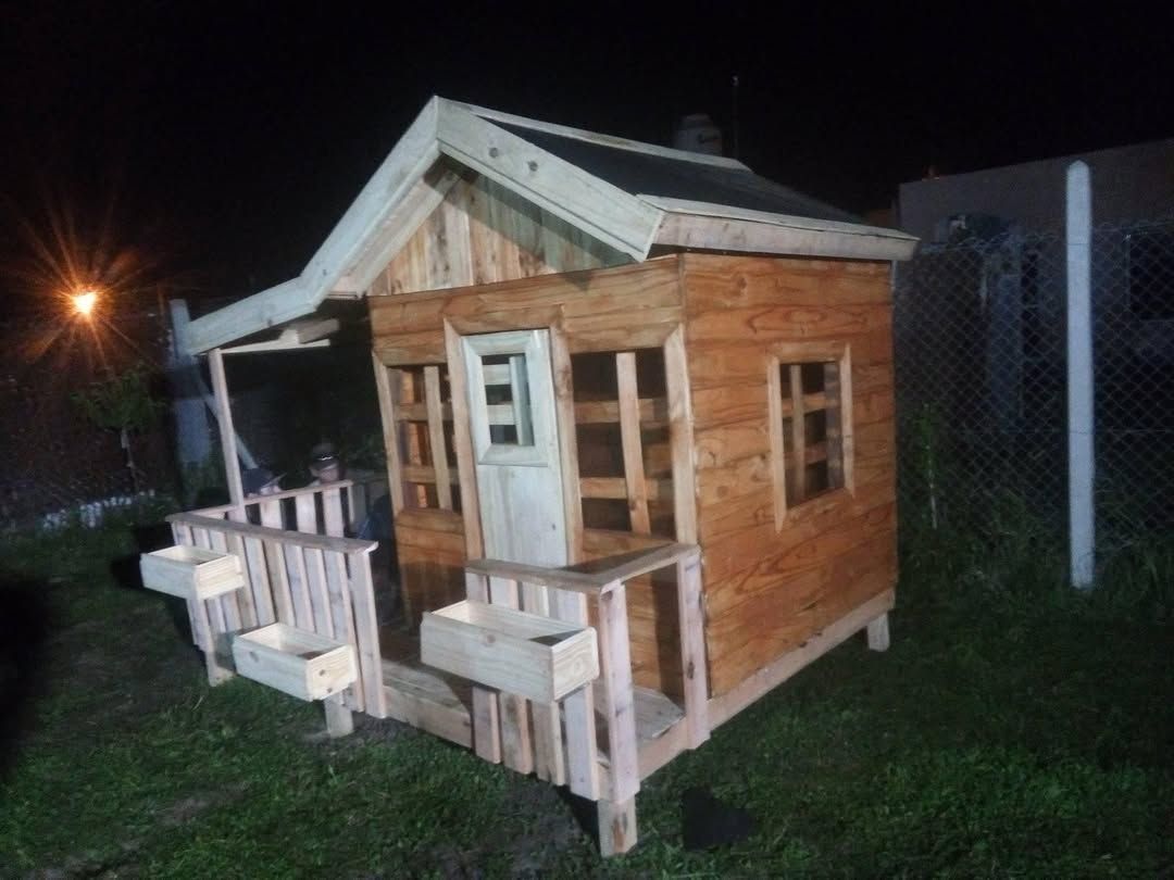 Una pequeña casita de madera con porche y barandillas, vista de noche en un jardín con césped.