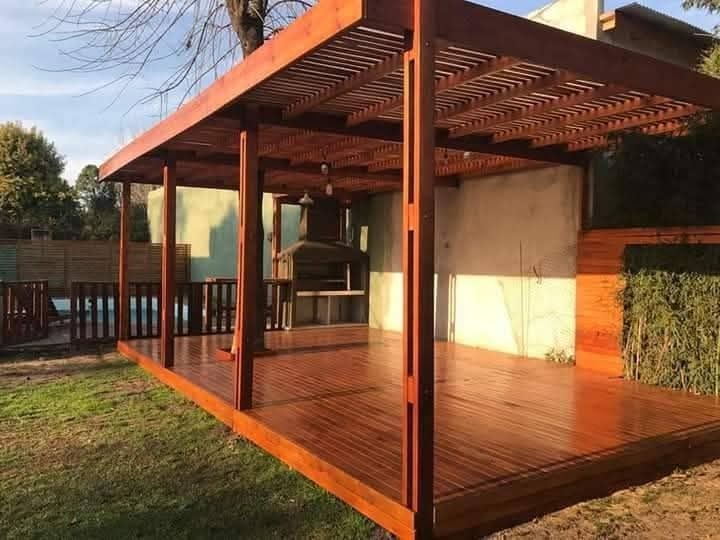 Patio cubierto con pérgola de madera y suelo pulido, junto a una piscina y un muro de seto en el jardín trasero.