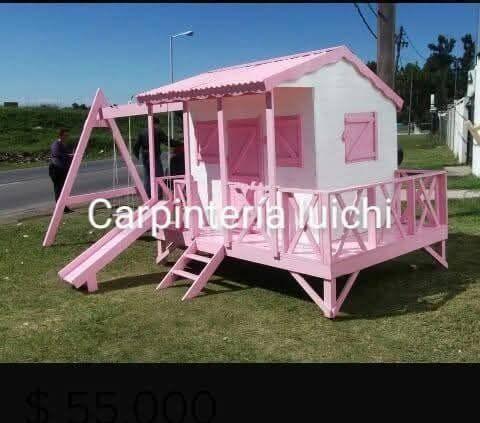 Minicasa rosa con porche, escaleras y columpio en un terreno cubierto de césped junto a la carretera.
