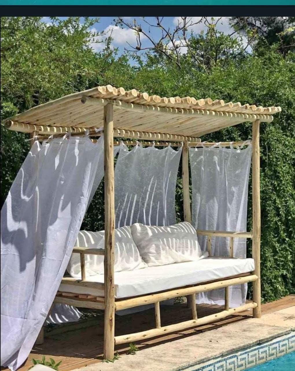 Cabaña de madera con diván y cortinas blancas junto a una piscina en un jardín frondoso.