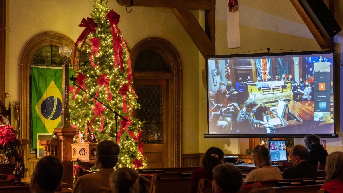 A group of people are sitting in front of a projector screen in a church with a christmas tree in the background.
