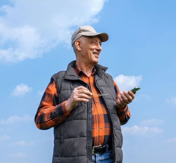 An older man is standing in a field holding a cell phone.