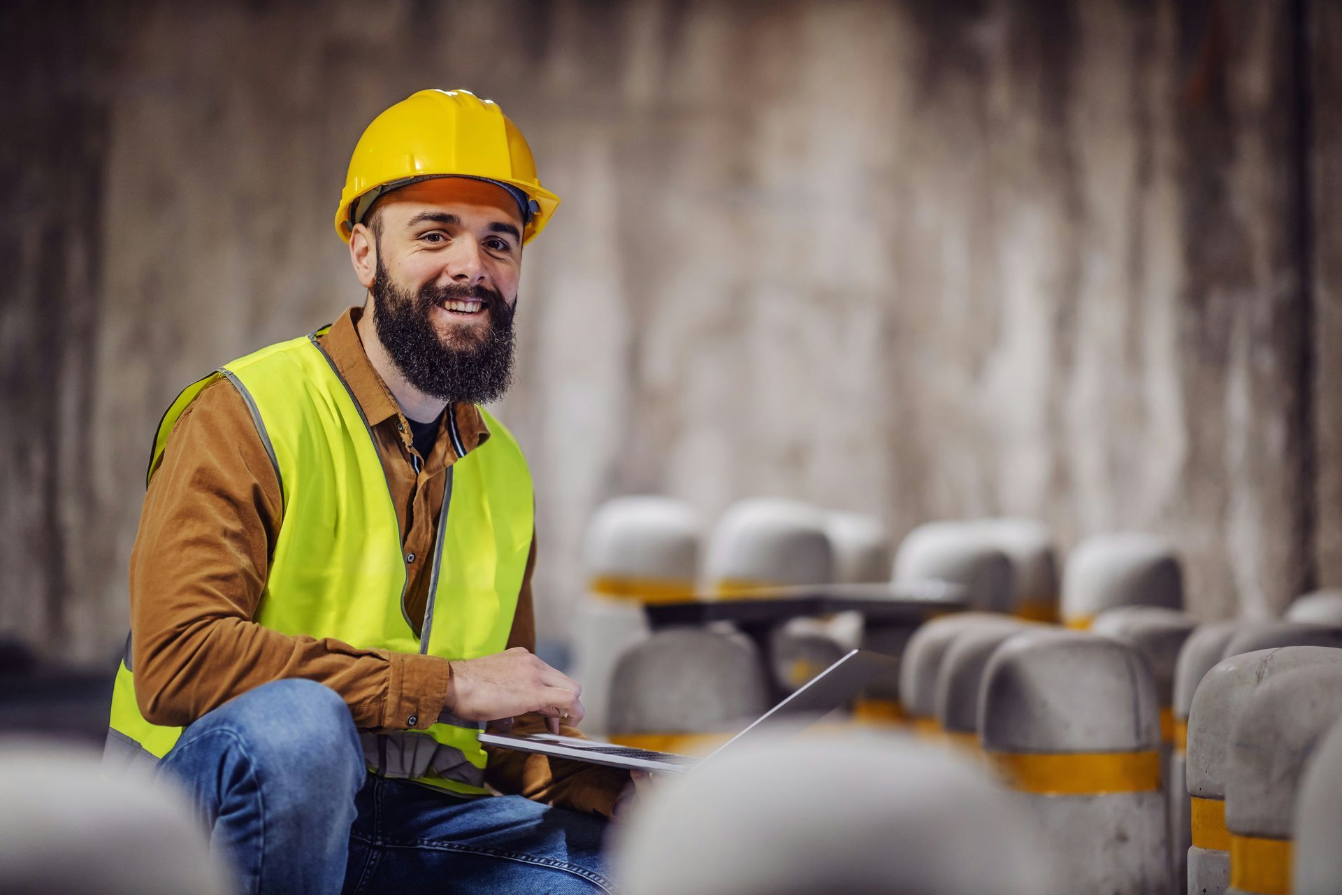 A man wearing a hard hat and safety vest is sitting in a warehouse holding a clipboard.