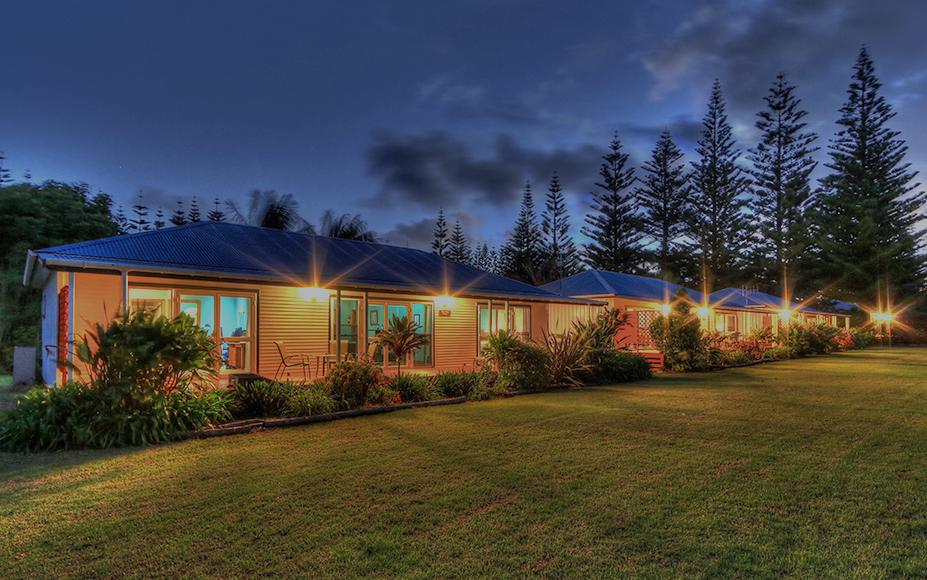Houses with lights on at dusk, green lawn, tall trees in background. Norfolk Island Trade Winds Seniors Sanctuary
