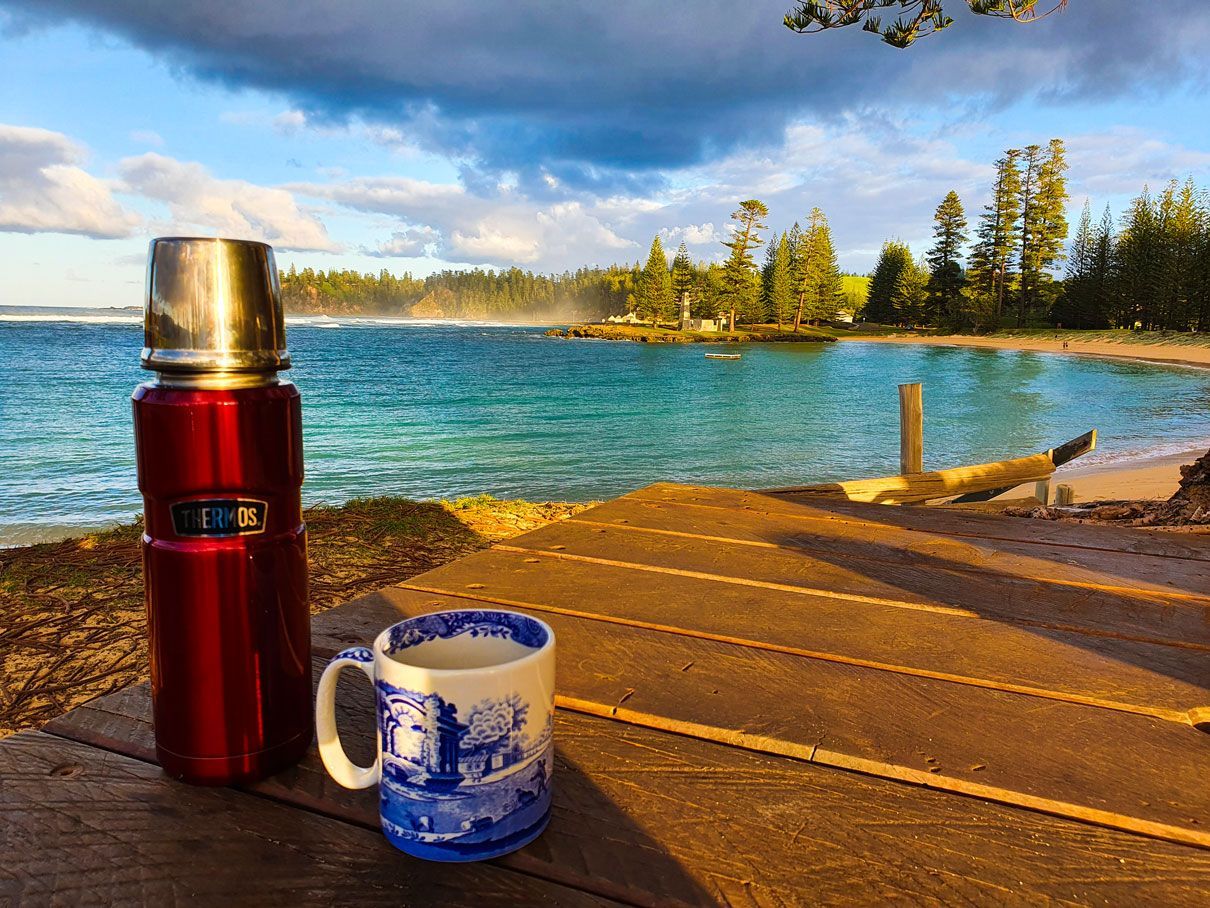 Red thermos and blue mug on a wooden table overlooking a turquoise beach. Norfolk Island Trade Winds Seniors Sanctuary