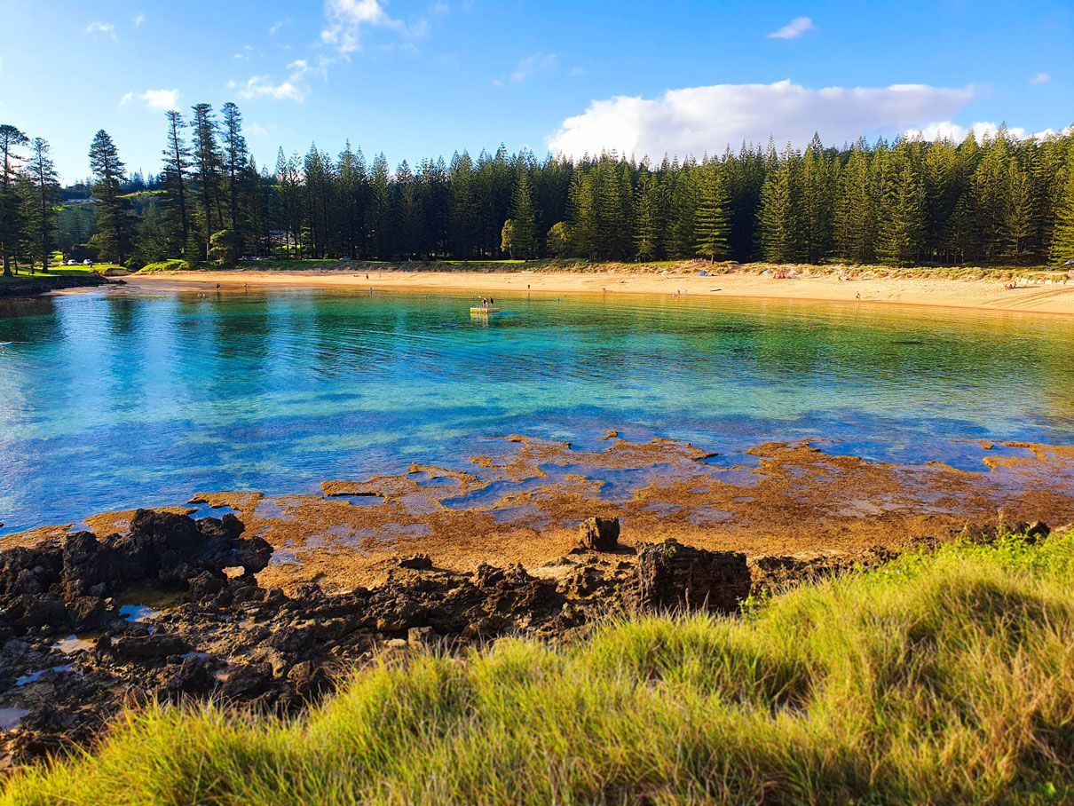 Blue and turquoise water with a sandy beach, framed by green trees. Norfolk Island Trade Winds Seniors Sanctuary