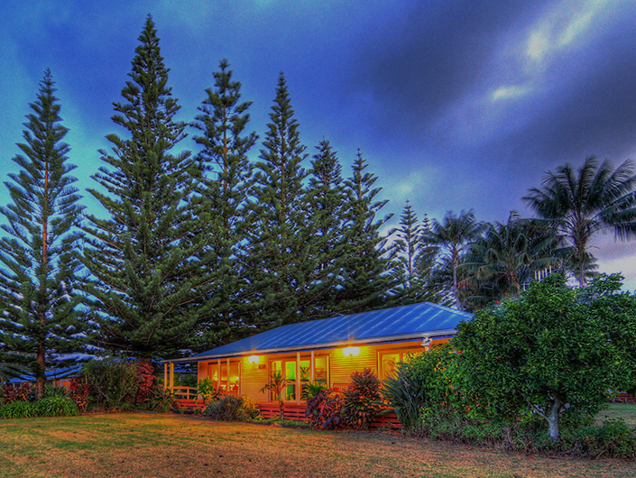 House lit up at dusk, tall pine trees, blue roof, lush greenery. Norfolk Island Trade Winds Seniors Sanctuary