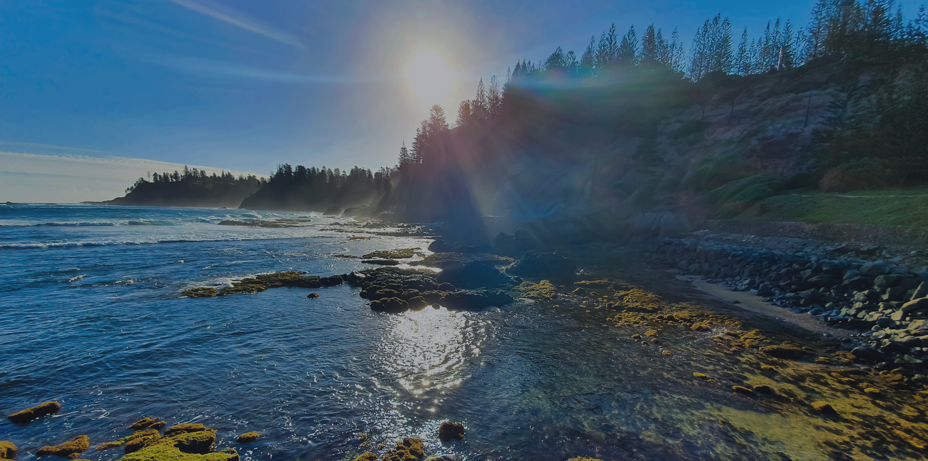 A sunny coastal scene with water, rocks, and trees on the shore. Norfolk Island Trade Winds Seniors Sanctuary