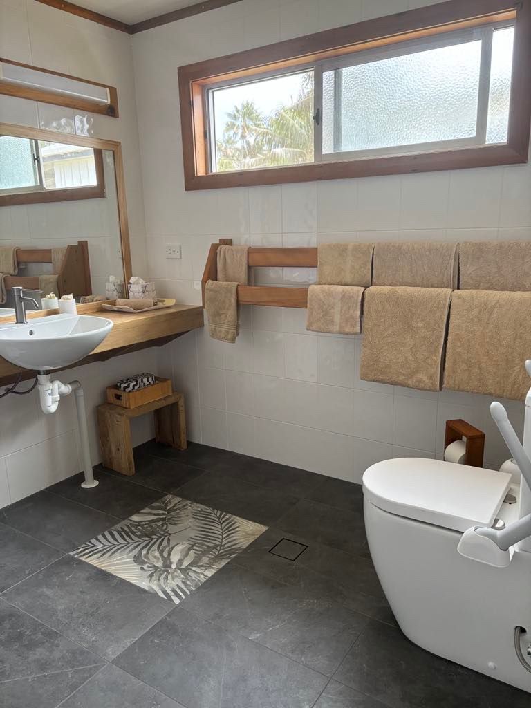 Bathroom with white walls, dark gray tiled floor, and wooden accents. Norfolk Island Trade Winds Seniors Sanctuary