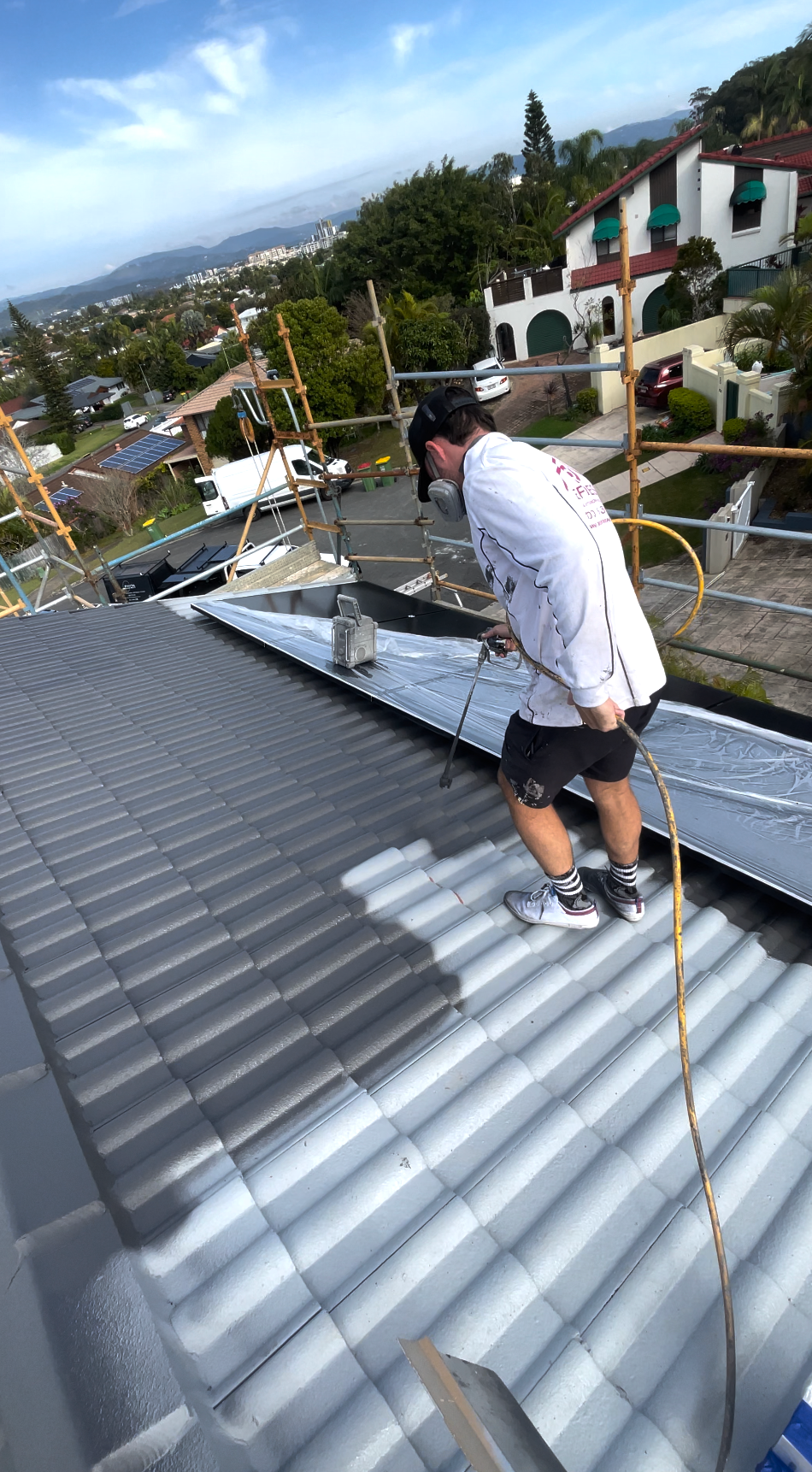 A person wearing a white long-sleeve shirt paints a grey roof while harnessed to a safety line on scaffolding.