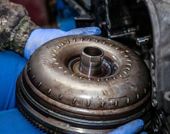 Person in blue gloves holds a vehicle torque converter, a circular metal component during repair.