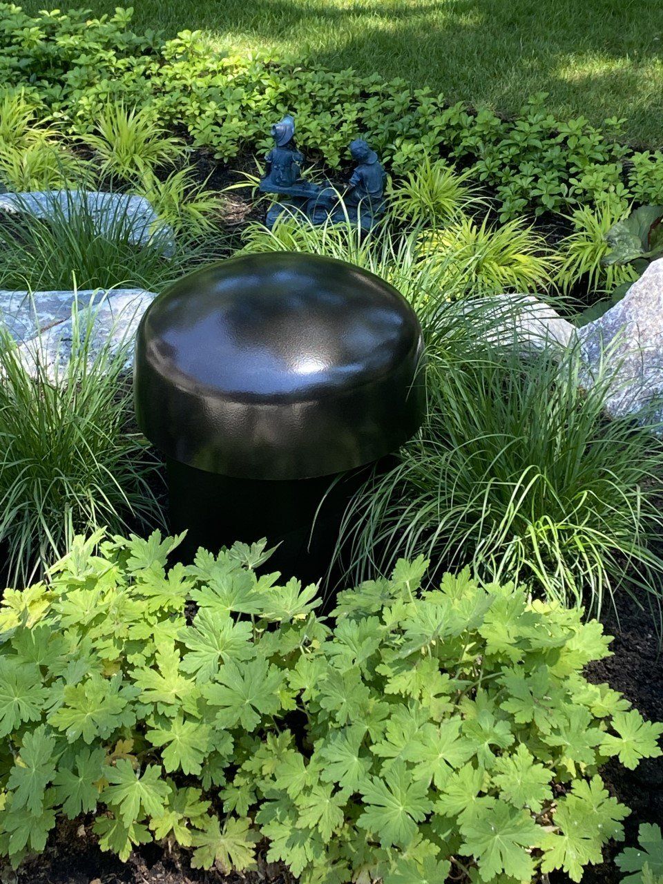 A black speaker is sitting in the middle of a garden surrounded by green plants.