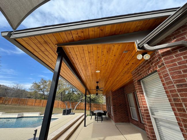 Covered patio with wood ceiling, black supports, brick house, pool, and blue sky.