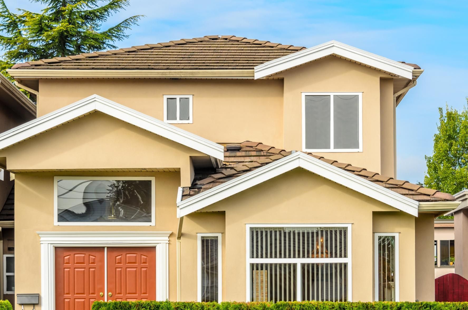 Two-story house with tan stucco exterior, brown roof, and white trim, with a double red front door and large windows.